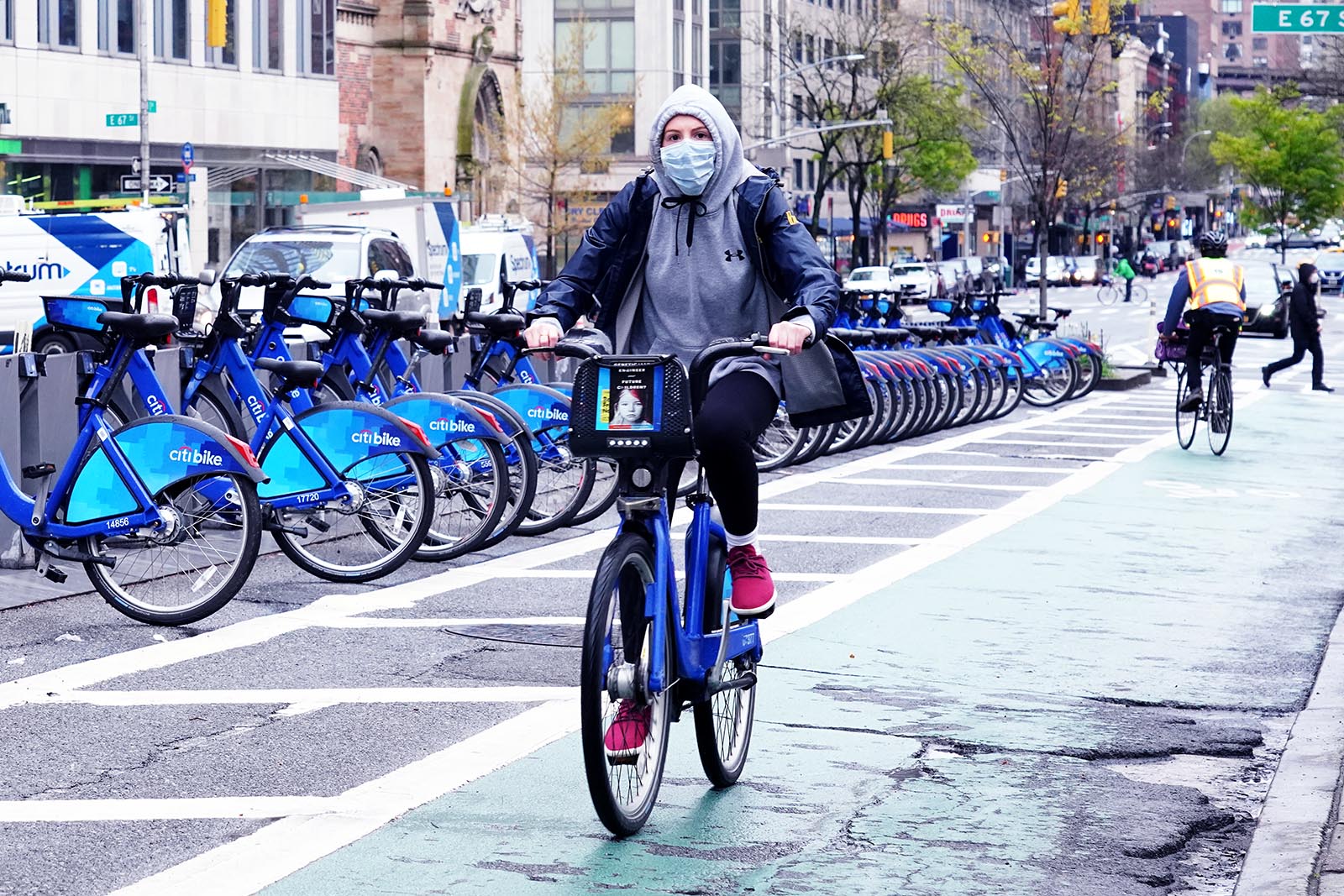 A woman rides a CitiBike in New York city while several other eBikes are parked in the background.